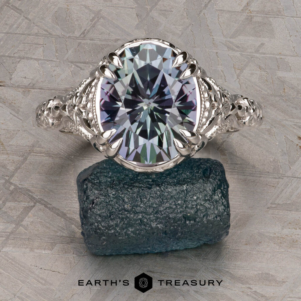 A silver ring from D'Amico, Amanda [custom gallery] with an oval-cut gemstone stands on a rough blue-green stone against a textured gray background, with "EARTH'S TREASURY" shown below.
