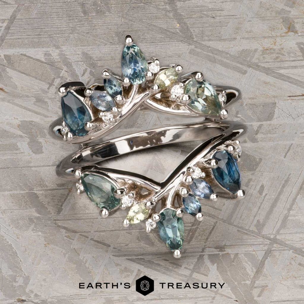 A custom "Brockman, Jeff and Young, Allene 2" silver ring with blue, green, and clear gemstones in a leaf-inspired design, shown on a textured gray surface with the "Earth's Treasury" logo at the bottom.