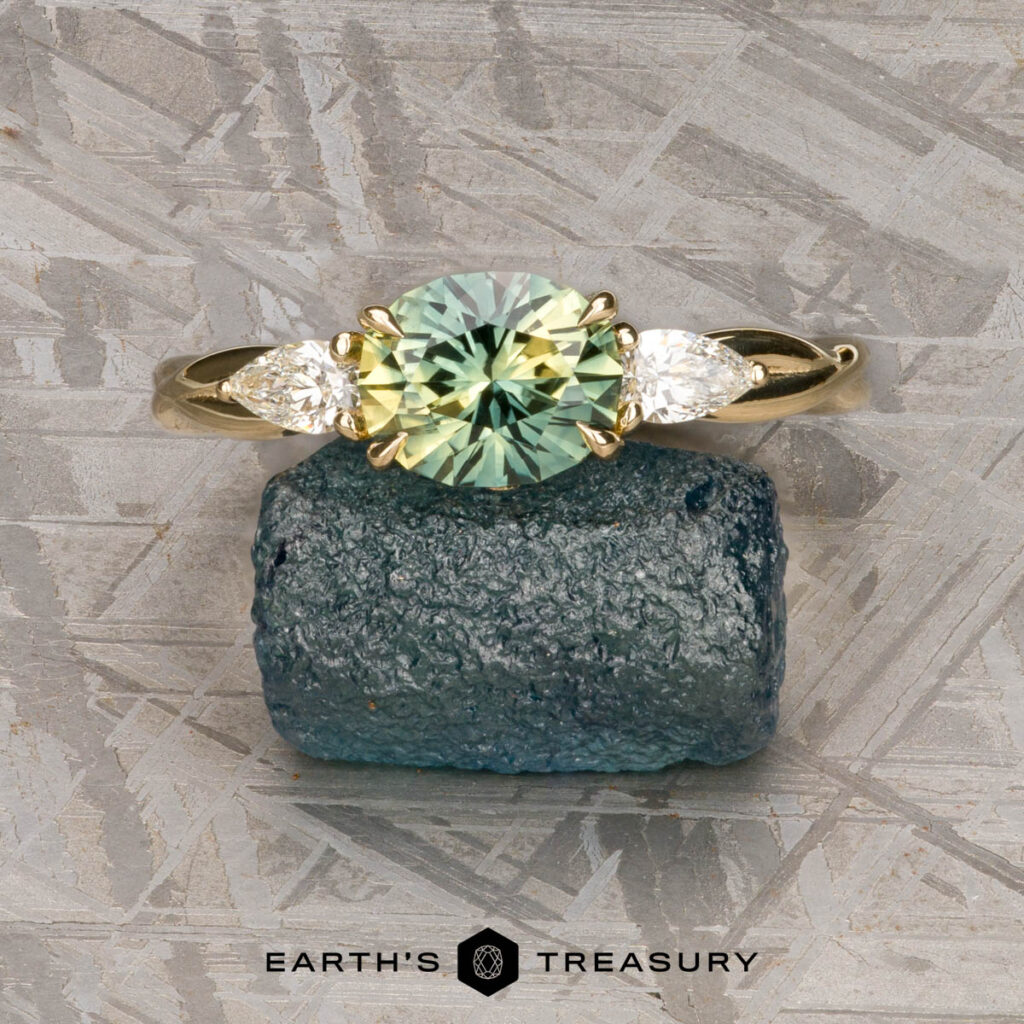 A gold ring from Burbridge, Mark [custom gallery], featuring a round green gemstone between two pear-shaped white stones, shown on a rough blue crystal with the "Earth's Treasury" logo beneath.