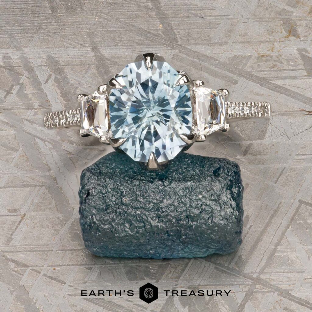 The Coll, Shawn 1 custom gallery ring features a faceted center stone with two side gems, displayed atop a rough blue-green gemstone, with the "Earth's Treasury" logo beneath.