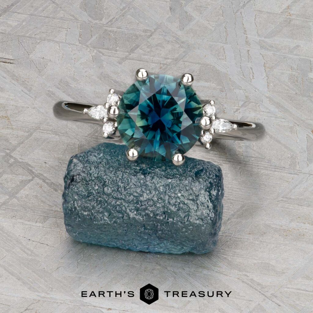 LeMoel, Jonathan 1 [custom gallery]: A silver ring with a round blue-green gemstone and small clear side stones is displayed on a rough blue stone, with "EARTH'S TREASURY" text beneath.