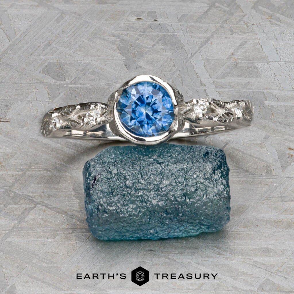 The Casseri, Eleni and Stevens, Matthew [custom gallery] features a silver ring with a round blue gemstone atop a rough blue crystal on a textured gray surface with "EARTH'S TREASURY" below.