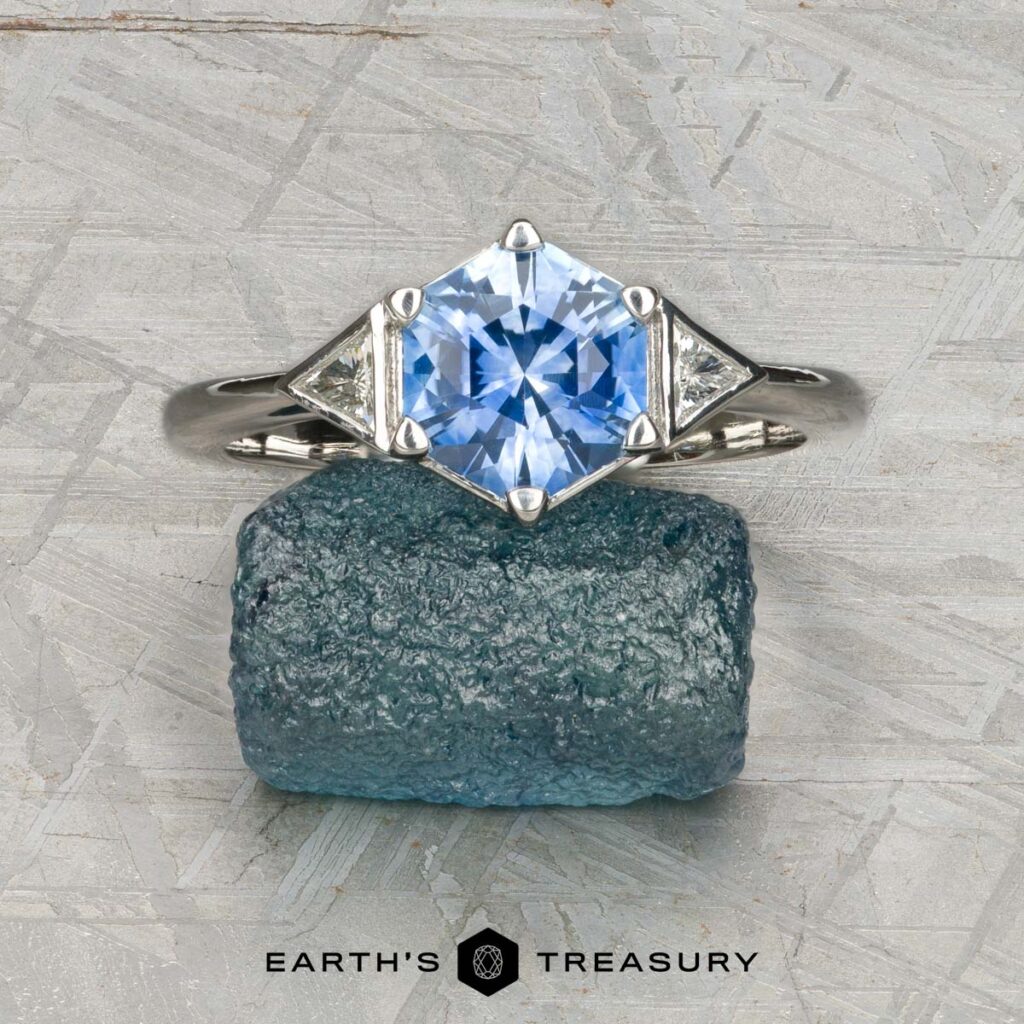 A silver ring named "Gabay, Alan" features a hexagonal blue gemstone and two triangular clear stones. It's displayed on a rough blue mineral with "Earth's Treasury" branding below.