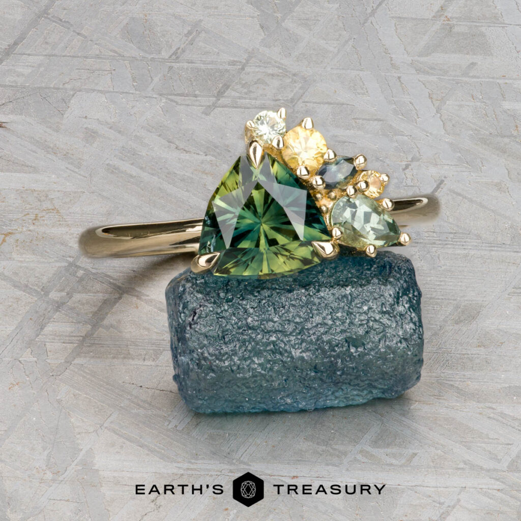 A custom gold ring by Locke, Jordan and LaGrua, Cayleigh features a large triangular green gemstone accented with smaller round yellow and light green stones, displayed on blue crystal. Logo: “Earth’s Treasury.”.