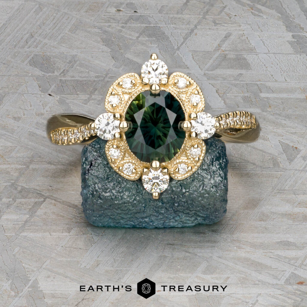 The Kuba, Steven [custom gallery] ring features a green oval gemstone encircled by diamonds, resting on a rough blue stone atop a textured gray surface. Earth’s Treasury logo appears below.