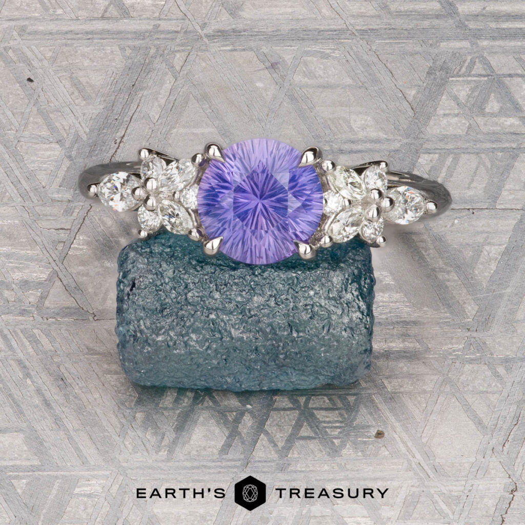 A silver ring featuring a round lavender gemstone and four clear side stones from Chu, Geoffrey and Lin, Iris [custom gallery] rests on a blue-green crystal, set against a geometric background with the "Earth's Treasury" logo.
