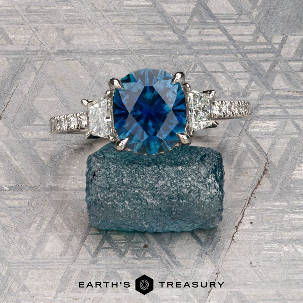 The "Martinez, Natalie 1 [custom gallery]" ring features an oval blue gemstone flanked by two clear side stones, displayed on a rough blue crystal atop a patterned gray surface, with "Earth's Treasury" written beneath.