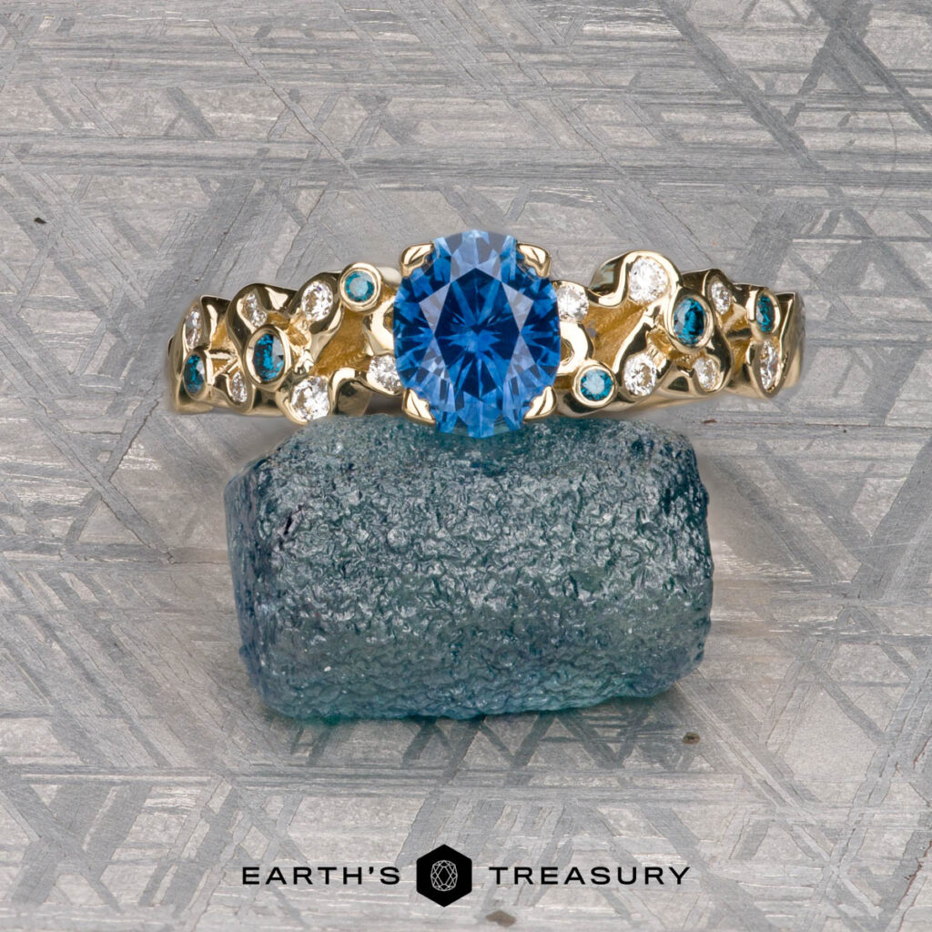 A gold ring from Contreras, Javier [custom gallery] with a blue gemstone and small diamonds is displayed on a blue textured stone against a patterned background. The "Earth's Treasury" logo appears below.