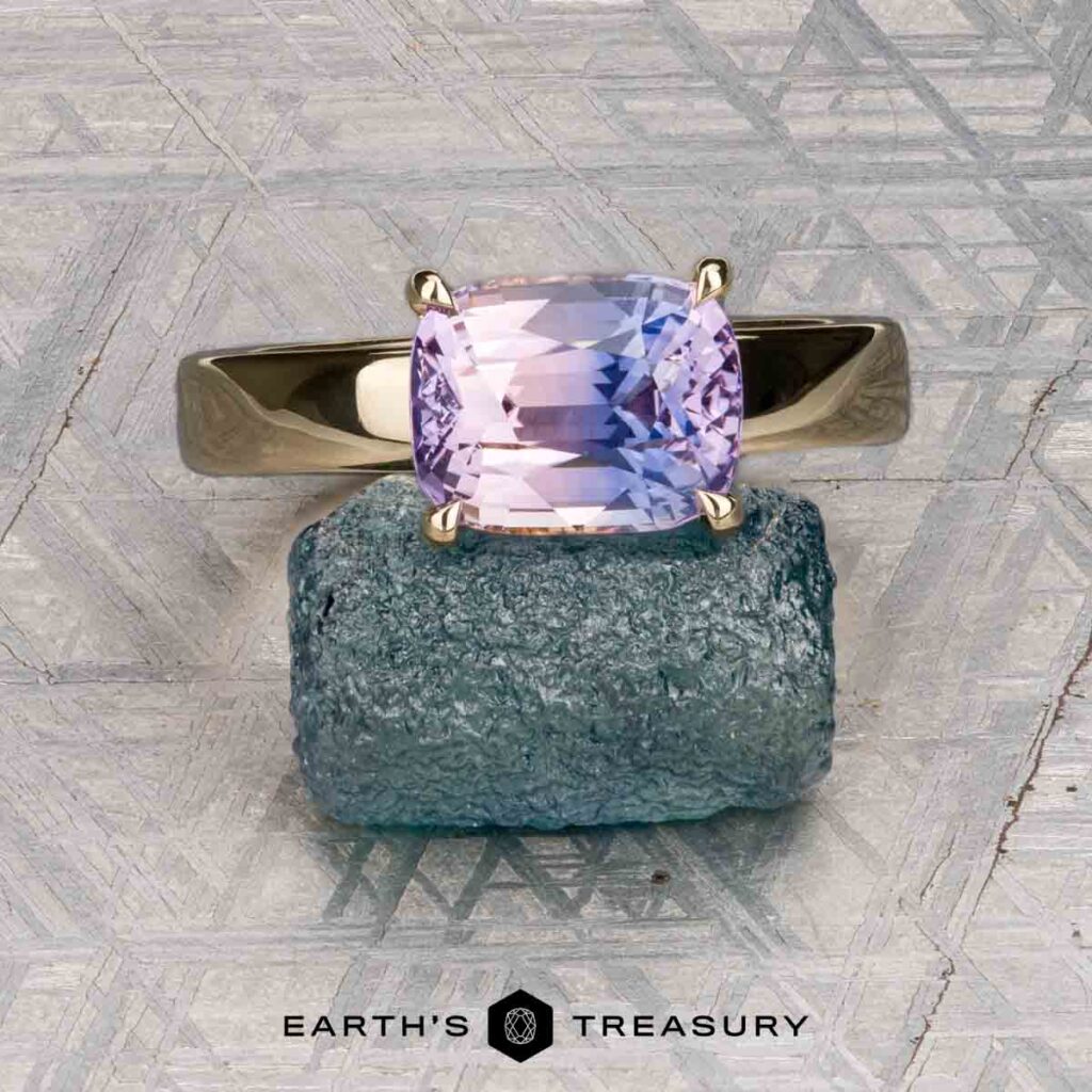 The Howe, Benjamin [custom gallery] gold ring with a cushion-cut purple gemstone rests on a rough blue-green stone, with the Earth's Treasury logo below.