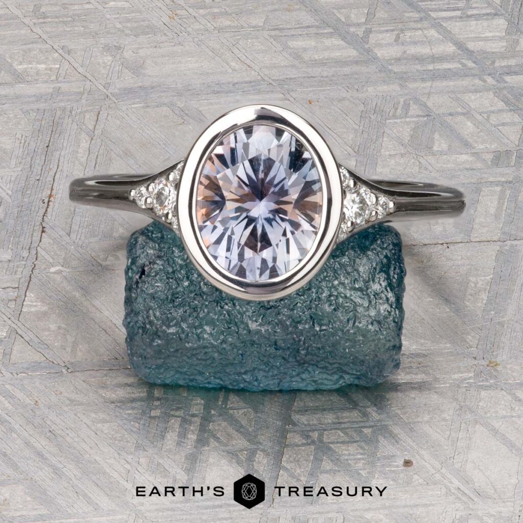 The Dobies, Jackson and Martin, Lizzie silver ring with an oval-cut gemstone and small side stones is displayed upright on a rough blue-green crystal, with the Earth's Treasury logo below.