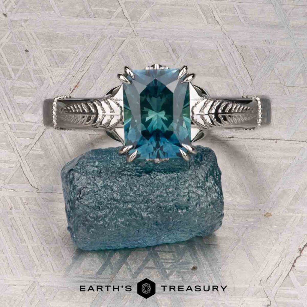 Jenkins, Casey and Wyatt, Damaris (Marit) 1 [custom gallery]: A blue-green faceted gemstone ring with leaf details rests on a rough blue crystal, set against a textured gray background.