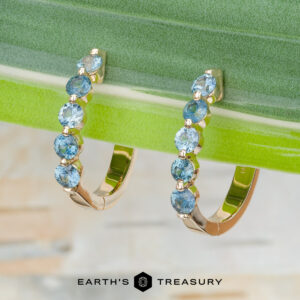 The "Eclipse" Earrings in 14K yellow gold—gold hoops adorned with six round blue gemstones each—are showcased against a green plant background, with the Earth’s Treasury logo displayed at the bottom.