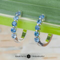 The "Eclipse" Earrings in 14K white gold: Pair of white gold hoop earrings set with round blue gemstones displayed on a green leaf, with the Earth's Treasury logo at the bottom.