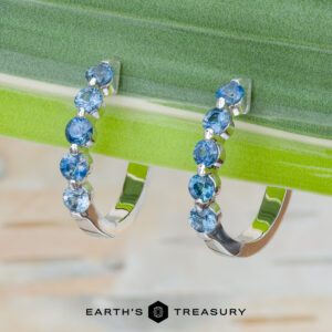 The "Eclipse" Earrings in 14K white gold: Pair of white gold hoop earrings set with round blue gemstones displayed on a green leaf, with the Earth's Treasury logo at the bottom.