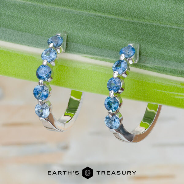 The "Eclipse" Earrings in 14K white gold The "Eclipse" Earrings in 14K white gold: Pair of white gold hoop earrings set with round blue gemstones displayed on a green leaf, with the Earth's Treasury logo at the bottom.