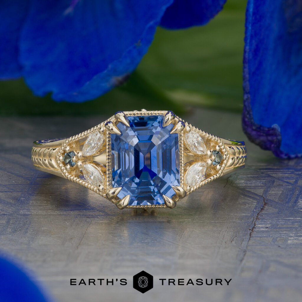 “Stacked from 20 images. Method=B (R=14,S=2): A gold ring with an emerald-cut blue gemstone, accented by marquise and round diamonds, displayed on a stone surface with blue flowers in the background.”.