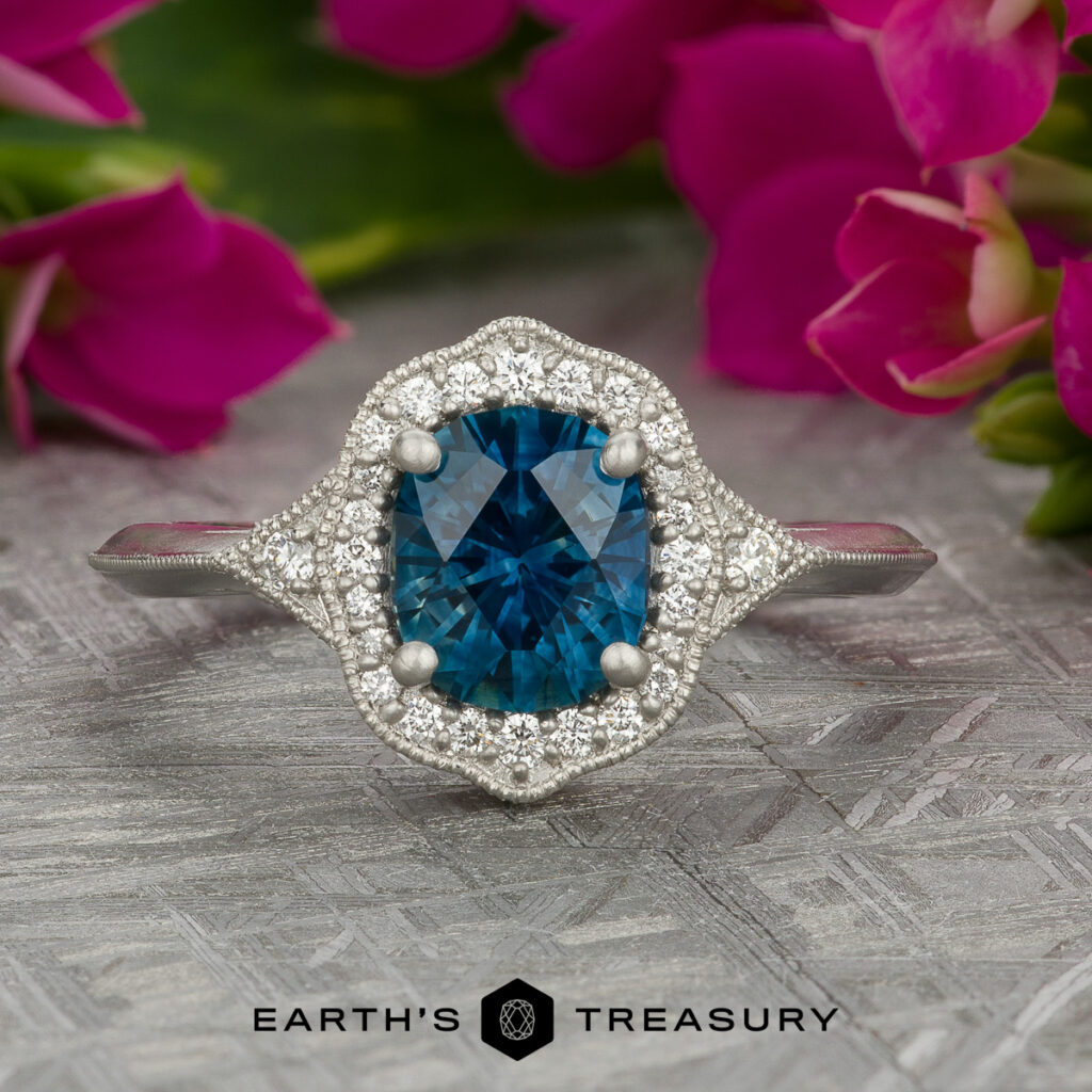 A silver ring featuring a cushion-cut blue gemstone and small white diamonds, shown on a textured surface with pink flowers and the "Earth's Treasury" logo. Product: "Stacked from 21 images. Method=B (R=7,S=2)".