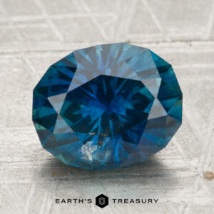 A 4.00-carat heated Montana sapphire, faceted in an oval shape, is displayed on a textured silver surface with the text "EARTH'S TREASURY" below.