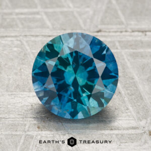 A round, faceted 2.29-carat Montana Sapphire (Heated) is displayed on a textured, light-colored surface with the text "EARTH'S TREASURY" at the bottom.