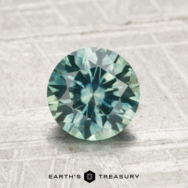 1.65-Carat Montana Sapphire (Heated) A 1.65-carat heated Montana sapphire, round and faceted in blue-green, stands upright on a textured light surface with the "EARTH'S TREASURY" logo below.