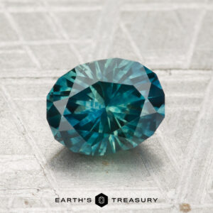 A 1.83-carat Montana Sapphire (Heated), round brilliant-cut and teal in color, is displayed on a textured light surface with "Earth’s Treasury" at the bottom.