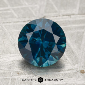 A 2.27-carat round brilliant-cut heated Montana sapphire is displayed on a textured light gray surface, with the "Earth's Treasury" logo and text at the bottom.