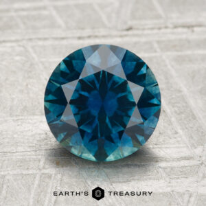 A 2.25-carat round brilliant-cut heated Montana Sapphire rests on a textured grey surface, with the "EARTH'S TREASURY" logo and text displayed at the bottom.