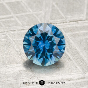 A 1.34-carat heated Montana sapphire, cut in a round faceted shape, rests on a textured metallic surface with "EARTH'S TREASURY" displayed beneath it.