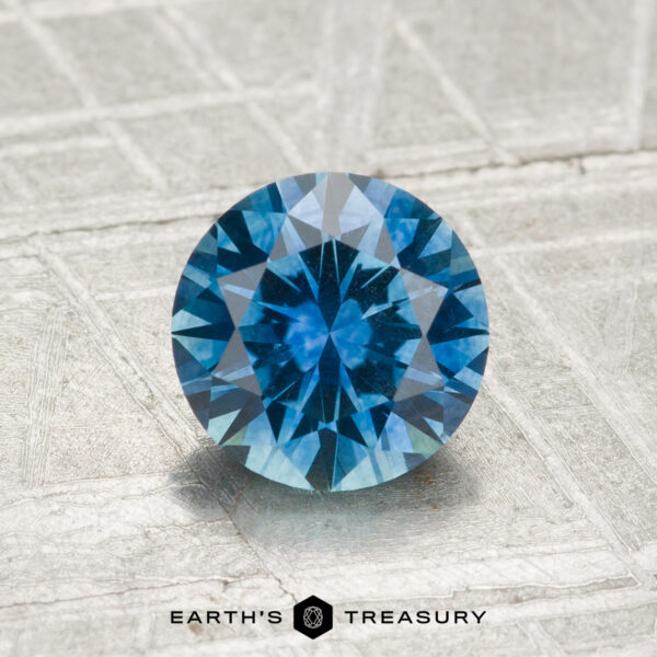 1.34-Carat Montana Sapphire (Heated) A 1.34-carat heated Montana sapphire, cut in a round faceted shape, rests on a textured metallic surface with "EARTH'S TREASURY" displayed beneath it.