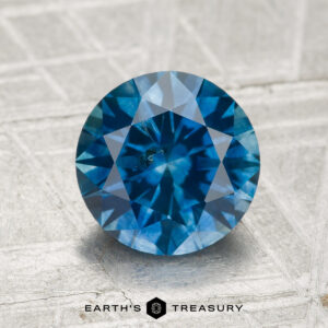 A 2.18-carat heated Montana sapphire with a round brilliant cut rests on a textured metallic surface, with "Earth's Treasury" displayed below.