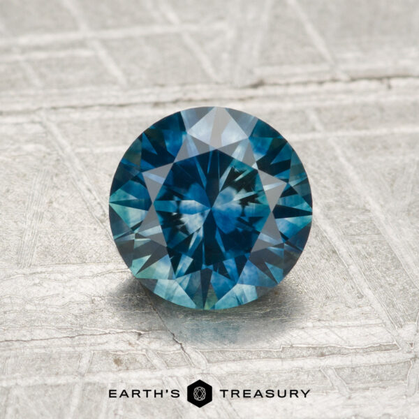 1.34-Carat Montana Sapphire (Heated) 1.34-Carat Montana Sapphire (Heated), round brilliant-cut blue gemstone, shown on a textured metallic surface with the "EARTH'S TREASURY" logo at the bottom.