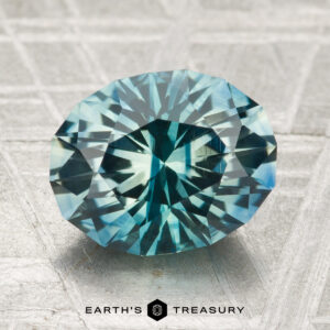 A 3.36-carat heated Montana sapphire, oval-cut with intricate facets in blue-green hues, is displayed on a textured metallic surface with "EARTH'S TREASURY" at the bottom.