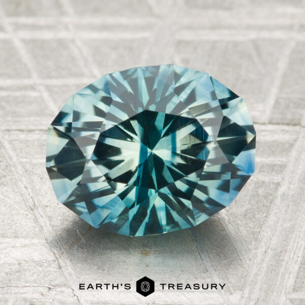 3.36-Carat Montana Sapphire (Heated) A 3.36-carat heated Montana sapphire, oval-cut with intricate facets in blue-green hues, is displayed on a textured metallic surface with "EARTH'S TREASURY" at the bottom.