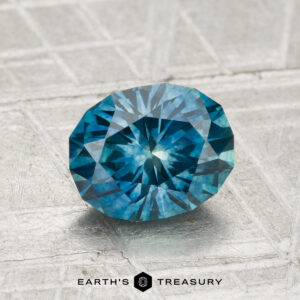 A 1.72-carat heated Montana sapphire with multiple facets is shown on a textured, light surface, with "EARTH'S TREASURY" and a hexagon logo beneath.