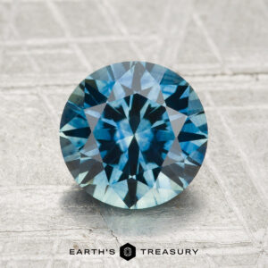 A 2.15-carat heated Montana sapphire in blue-green, round brilliant-cut, shown on a textured background with "EARTH'S TREASURY" at the bottom.