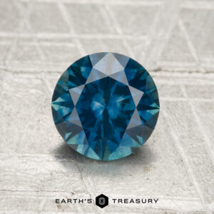 A 1.33-carat heated Montana sapphire, round brilliant-cut and blue in color, shown on a textured surface with "Earth's Treasury" beneath the stone.