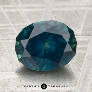 A 3.21-carat oval-cut heated Montana sapphire in deep blue rests on a textured surface, with "EARTH'S TREASURY" and a logo displayed below.