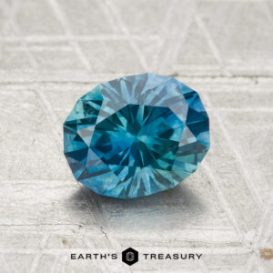A 1.70-carat heated Montana sapphire with a faceted blue surface rests on textured gray, "EARTH'S TREASURY" logo and text displayed beneath the stone.