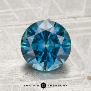 A 2.15-carat heated Montana Sapphire in a round brilliant cut is shown on a textured surface, with "EARTH'S TREASURY" displayed below.