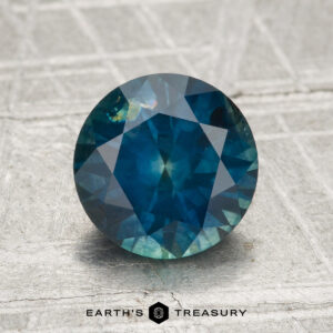 A 2.11-carat heated Montana sapphire, round and faceted in blue-green tones, rests on a textured metallic surface with "Earth's Treasury" displayed at the bottom.