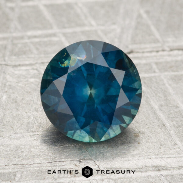2.11-Carat Montana Sapphire (Heated) A 2.11-carat heated Montana sapphire, round and faceted in blue-green tones, rests on a textured metallic surface with "Earth's Treasury" displayed at the bottom.