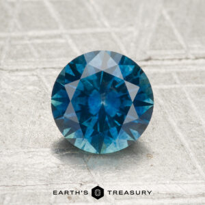 A 1.31-carat heated Montana sapphire in a round brilliant cut rests on a textured light gray surface, with "Earth's Treasury" written below.