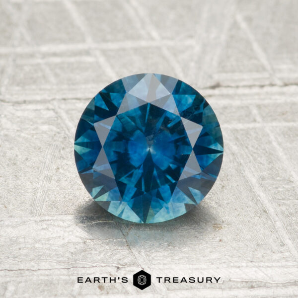 1.31-Carat Montana Sapphire (Heated) A 1.31-carat heated Montana sapphire in a round brilliant cut rests on a textured light gray surface, with "Earth's Treasury" written below.