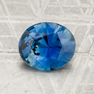 A 2.08-carat round, faceted blue sapphire is displayed on a textured gray surface with the "Earth's Treasury" logo at the bottom.