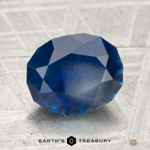 A 3.06-carat heated Montana sapphire with an oval cut is showcased on a textured metallic surface, featuring the "Earth's Treasury" logo at the bottom.