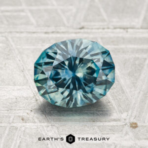 A 1.69-carat heated Montana sapphire, showcasing an oval cut and blue-green hue, rests on a textured metallic surface with "EARTH'S TREASURY" at the bottom.