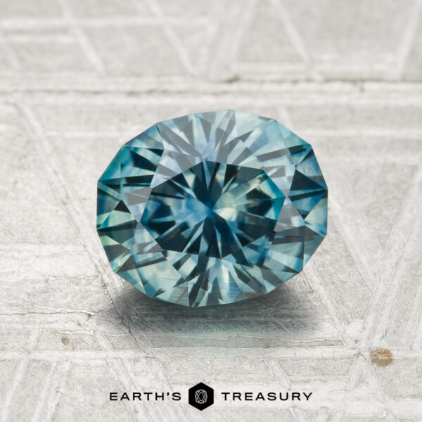 1.69-Carat Montana Sapphire (Heated) A 1.69-carat heated Montana sapphire, showcasing an oval cut and blue-green hue, rests on a textured metallic surface with "EARTH'S TREASURY" at the bottom.