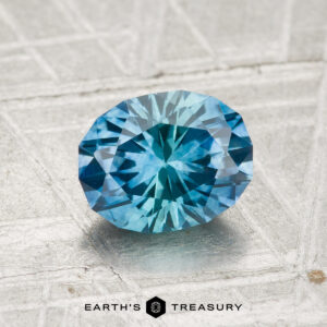 A 1.55-carat, oval, faceted Montana sapphire (heated) is showcased on a textured light gray surface with "EARTH'S TREASURY" displayed at the bottom.