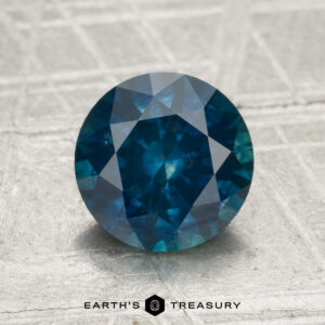 A 2.06-carat heated Montana sapphire, round-cut and blue, is shown on a light textured surface with "EARTH'S TREASURY" displayed at the bottom.