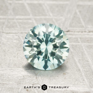A 1.29-carat heated Montana sapphire with a round, faceted cut and pale blue-green hue is displayed on a textured light gray surface. "EARTH'S TREASURY" appears at the bottom.